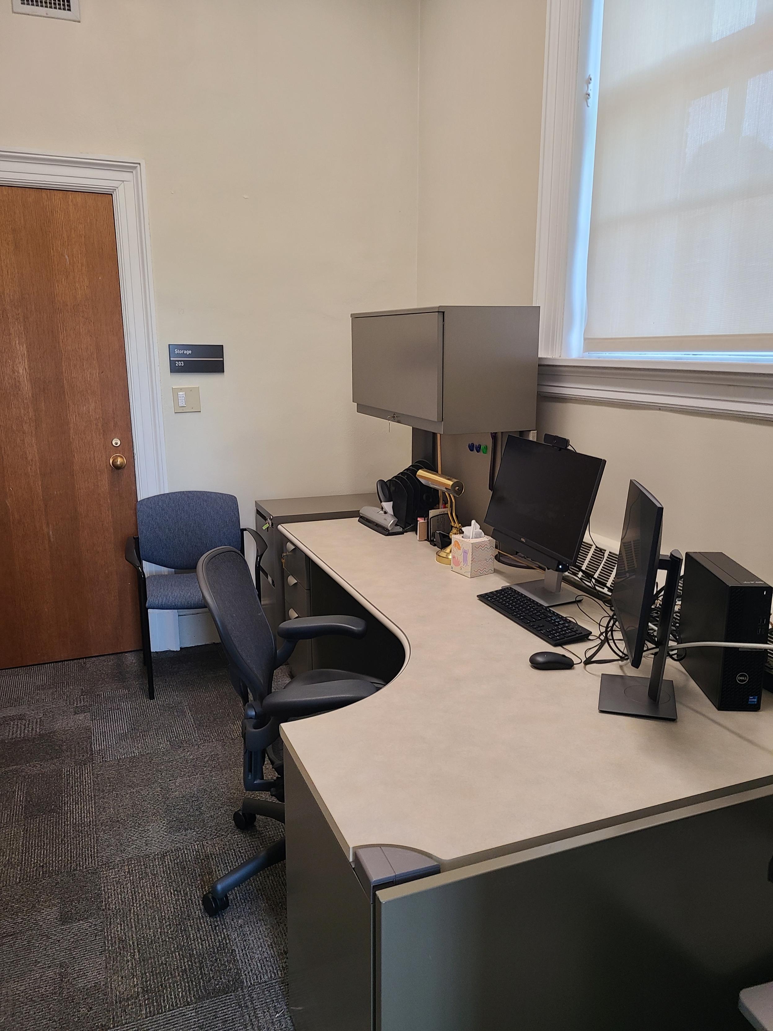 Office with a large corner desk facing a large window with the shade drawn, a PC workstation with two monitors, a small desk lamp and overhead enclosed bookshelf, a comfortable chair and second side chair.