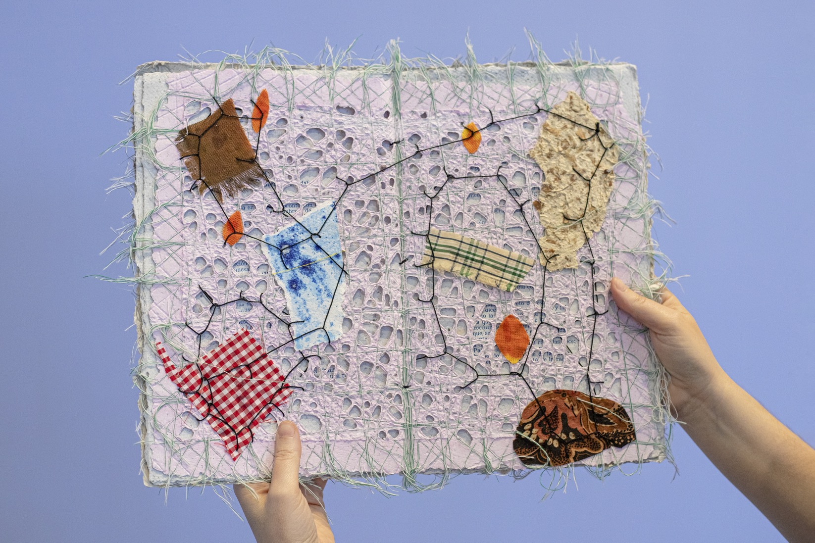 Handmade book of fabric and paper with many holes, wild stitching and fabric remnants. Held up by two hands against a plain light blue background.