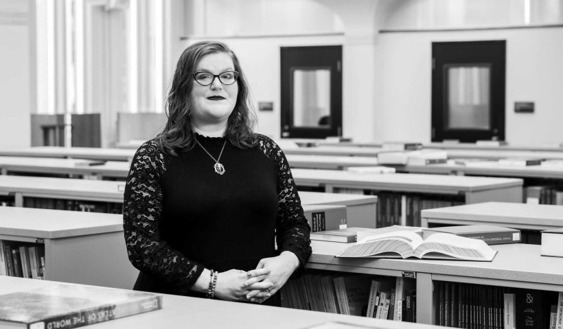 A woman, in black and white, wearing glasses and a black dress, standing in between shelves of books.