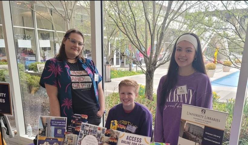 Three students in purple stand behind a UAlbany Libraries table in front of the fountain.