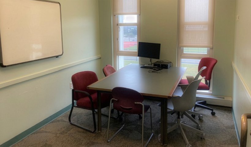 Table with 5 chairs around it sits in an empty room with windows, a whiteboard on one wall and an open door.
