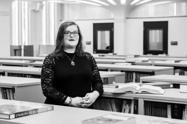 A woman, in black and white, wearing glasses and a black dress, standing in between shelves of books.
