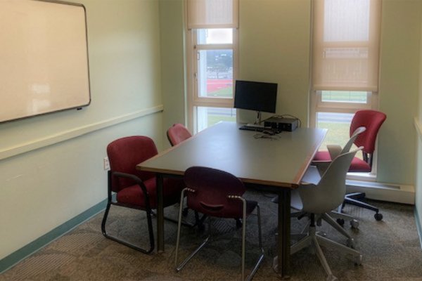 Table with 5 chairs around it sits in an empty room with windows, a whiteboard on one wall and an open door.