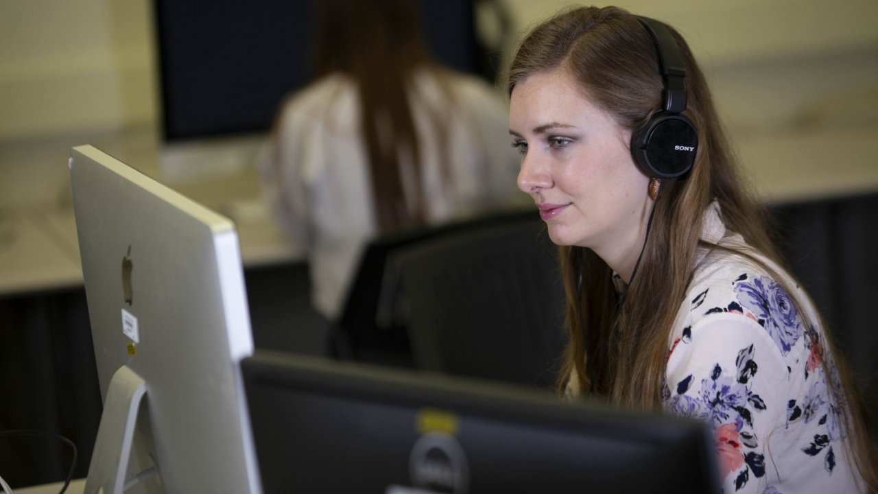 Student with long hair wearing headphones smiles while working at a Mac.