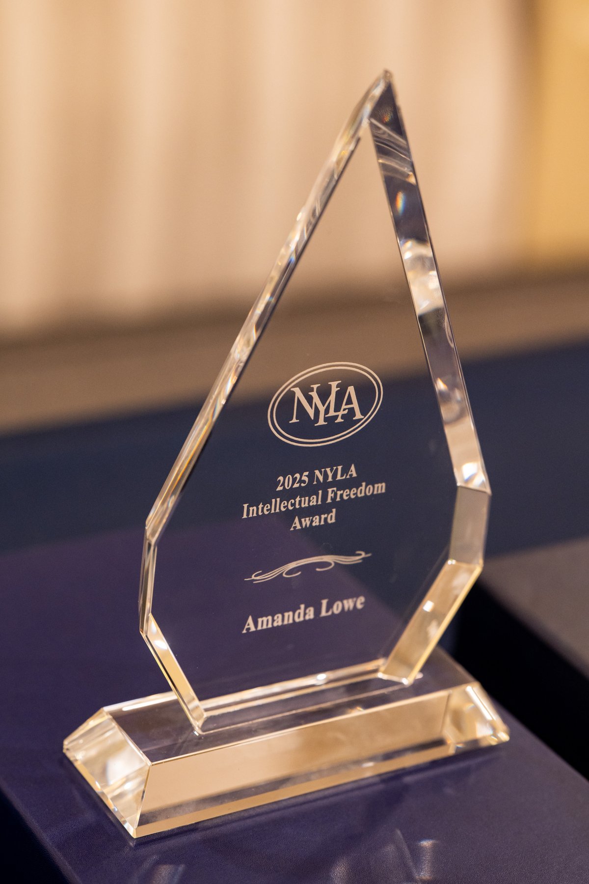A glass, triangular award with the text "2025 NYLA Intellectual Freedom Award: Amanda Lowe"