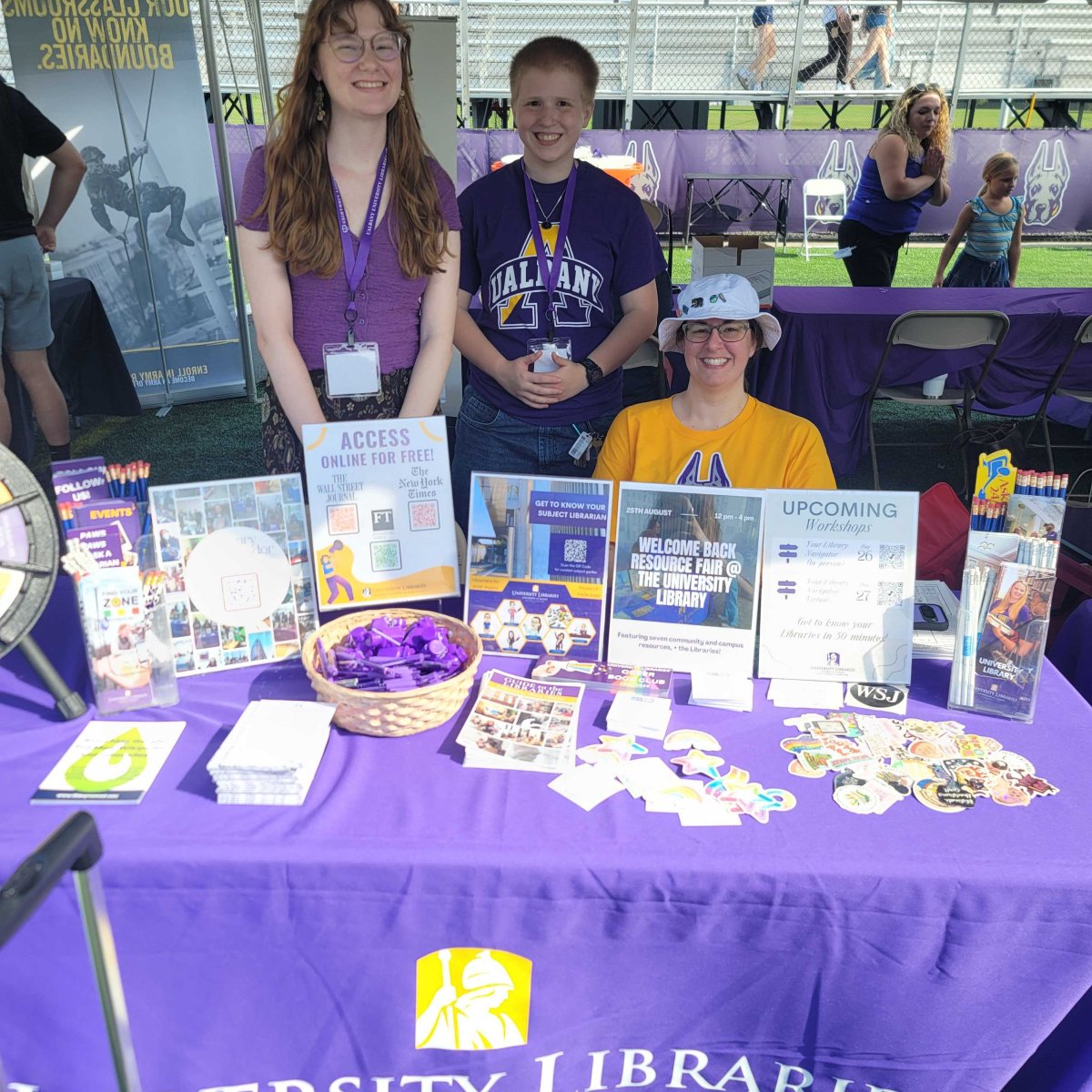 Two students in purple shirts and one staff member in gold shirt at a library event table