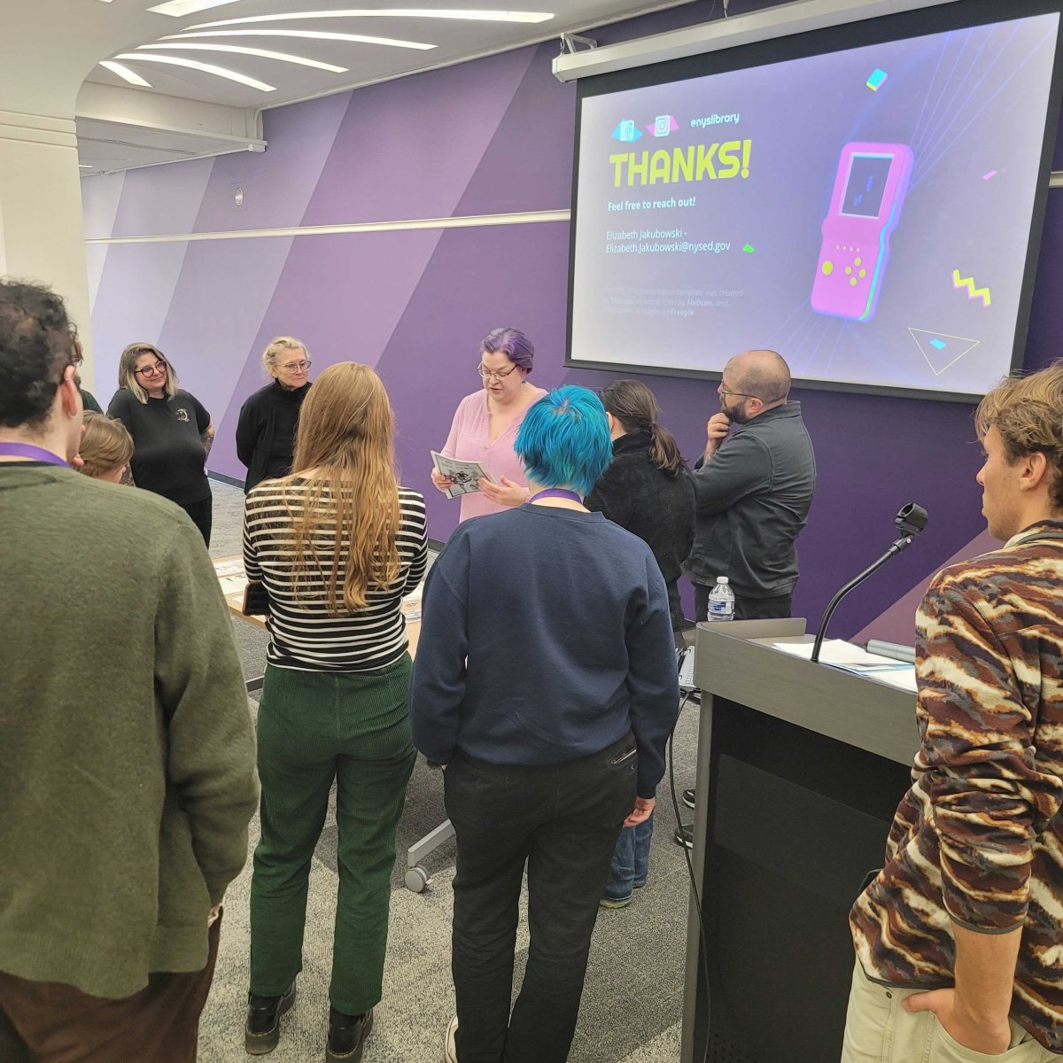 A woman in a pink shirt delivering a presentation in a semi-circle of attendees.