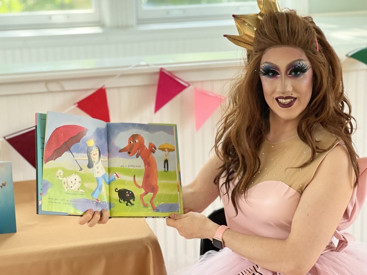 A Drag queen wearing a brown wig and pink dress smiles for the camera while holding an open children's book to an illustration of a dog dancing with a child