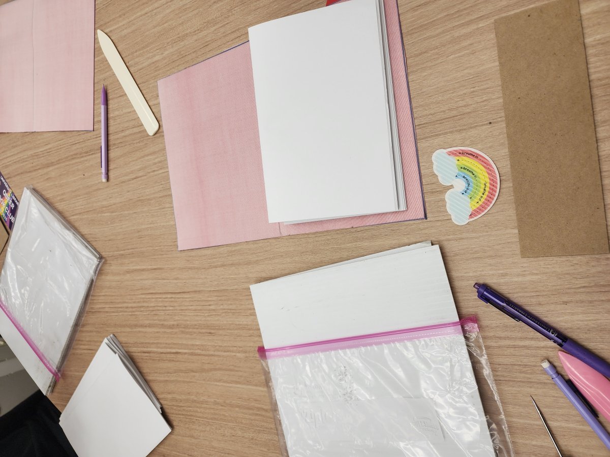 A desk space with white and pink paper and book-binding materials, along with a rainbow sticker