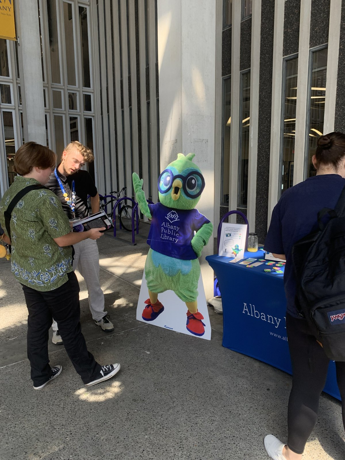 Two individuals reviewing a book next to a cut-out of Bleecker the APL mascot