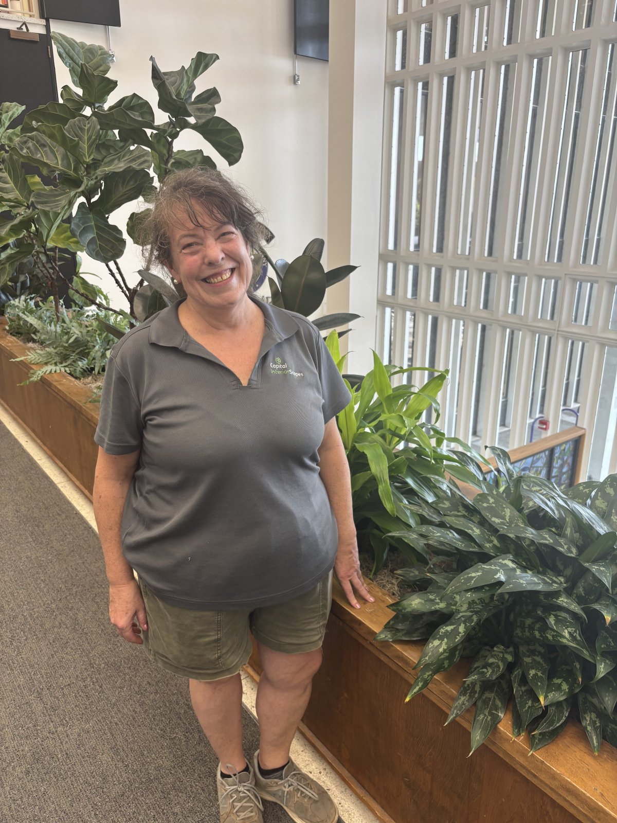 A woman in a gray polo shirt poses in front of a row of plants in the University Library