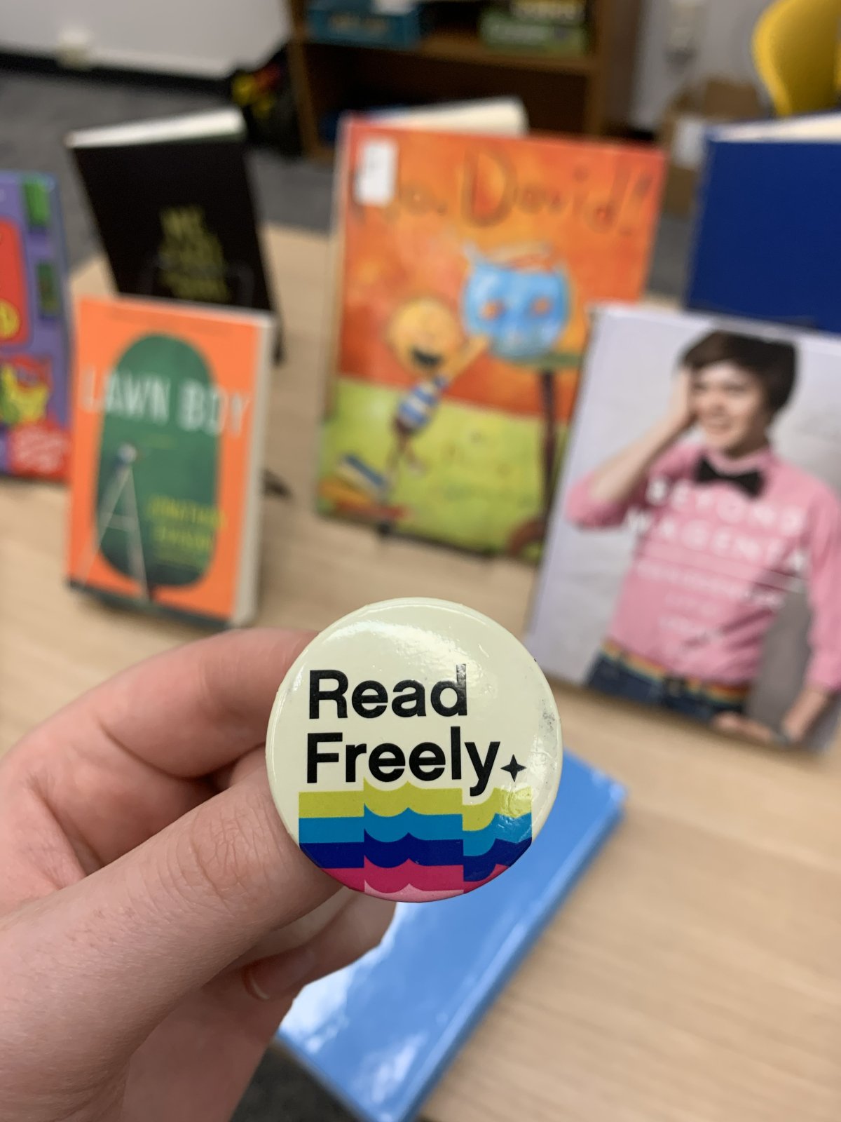 A hand holding a "Read Freely" button in the foreground with a table of banned books in the background.