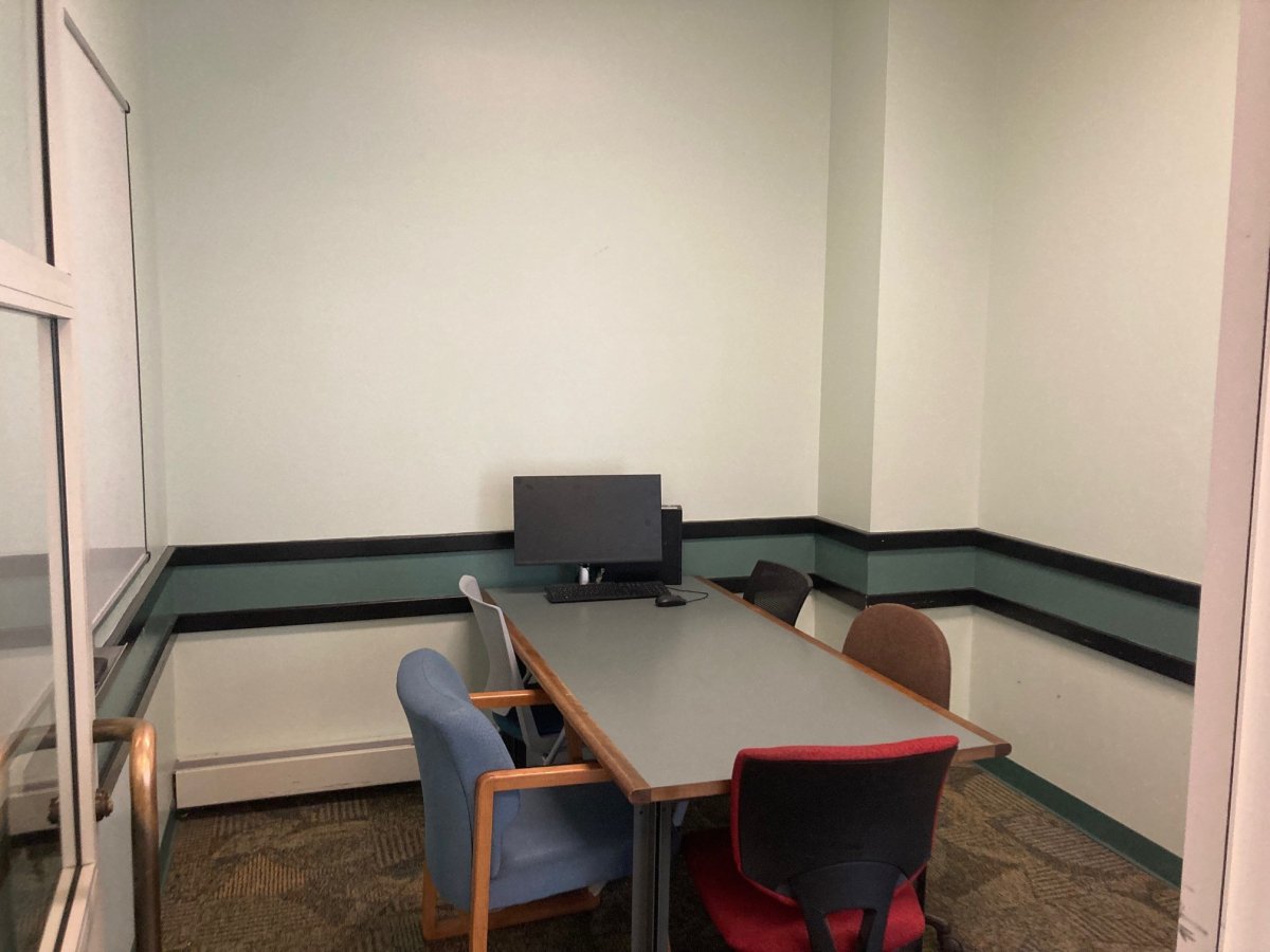 Table with a computer and 5 chairs around it sits in an empty room with a whiteboard on one wall and an open door.