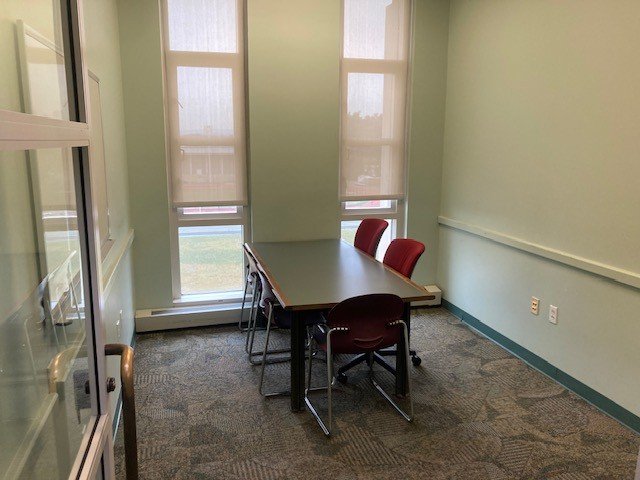 Table with 5 chairs around it sits in an empty room with windows, a whiteboard on one wall and an open door.