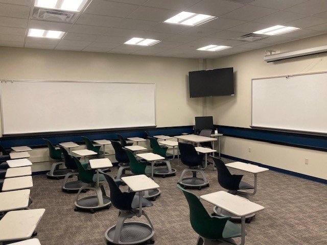 Bright classroom with whiteboards mounted on two walls, a television monitor mounted in the corner connected to the computer below it. Several hybrid desk/chairs on round, rolling bases are spread around the room.