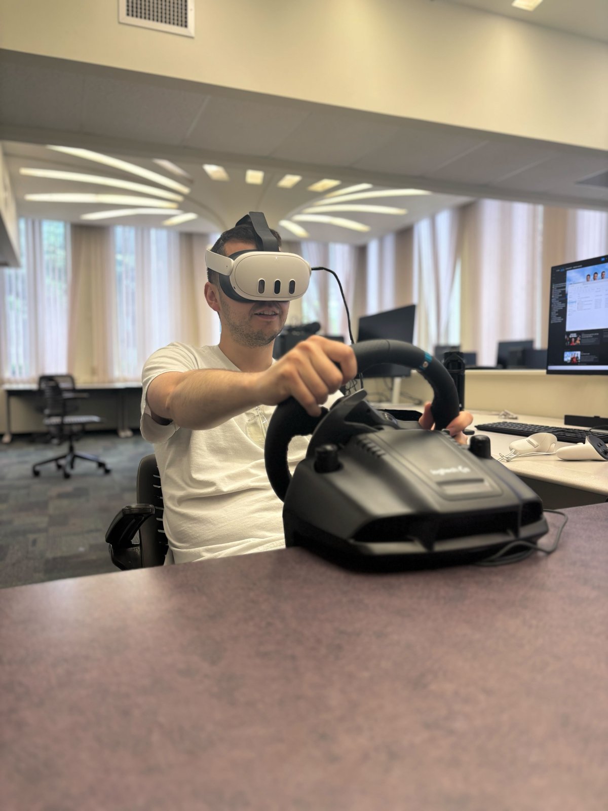 A student in a white shirt and wearing a white VR headset uses a video game steering wheel controller.