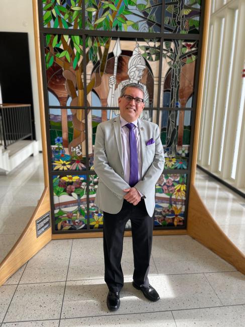 Dr. Gilbert Valverde posing in front of a stained-glass portrait of Minerva