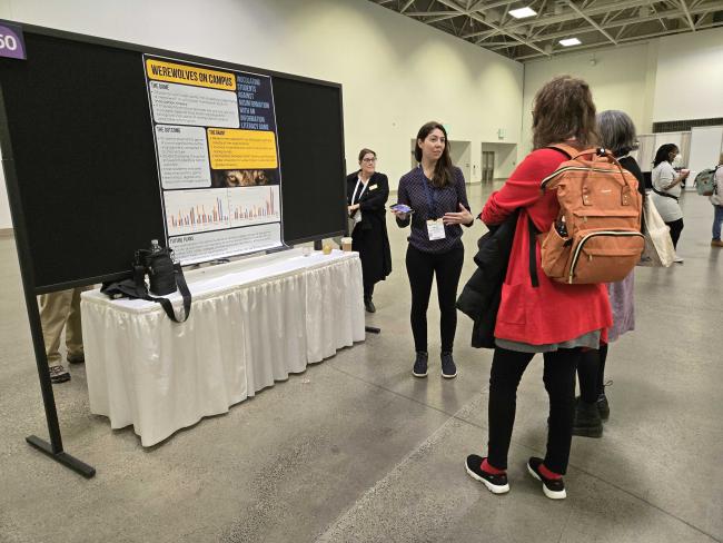 Kelsey O'Brien presenting a poster titled Werewolf on Campus to a group of conference attendees.