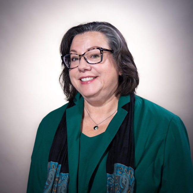 Headshot of woman smiling wearing funky glasses and a teal suit with black scarf, with chin-length, salt-and-pepper hair.